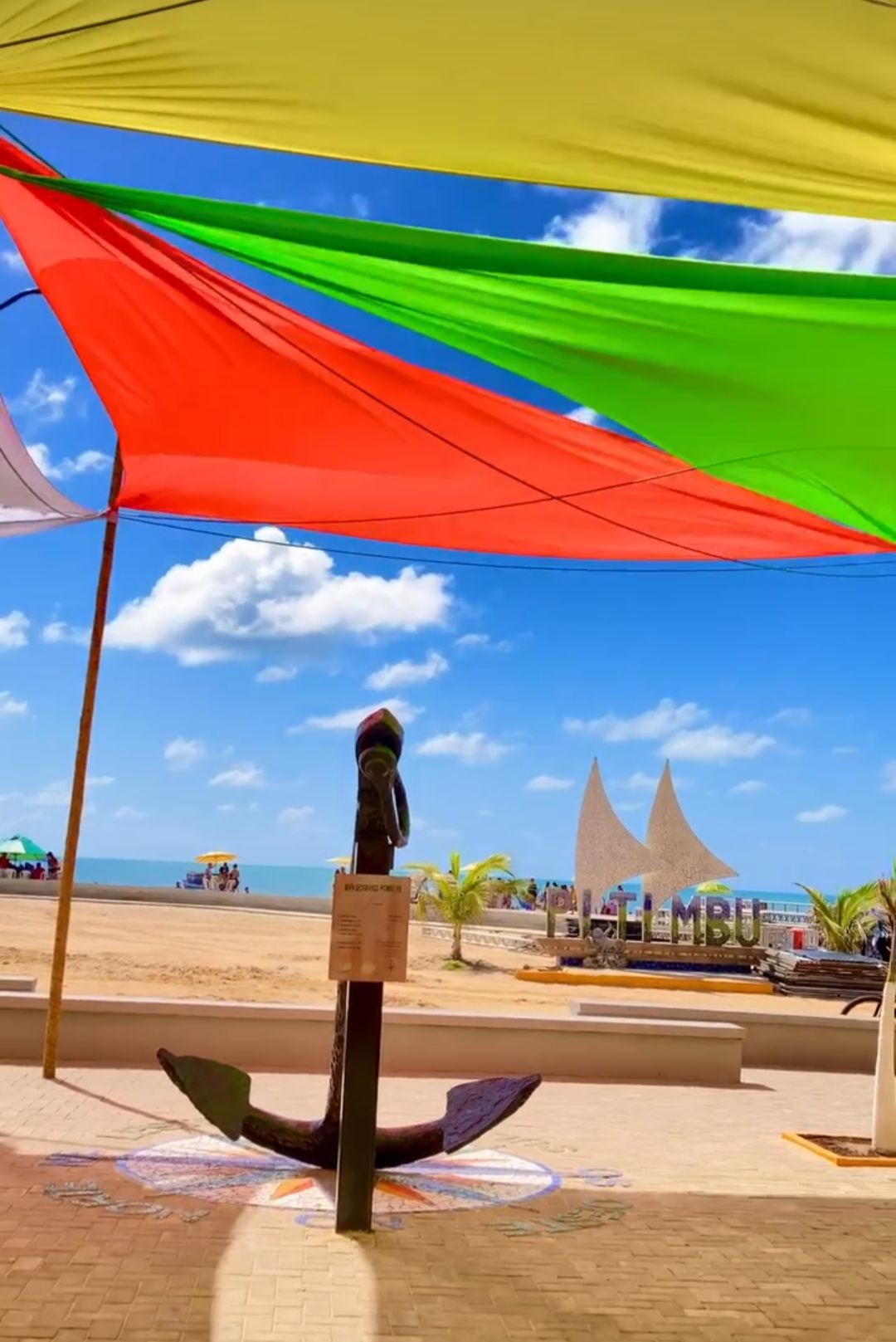 A vibrant view of the beach is framed by colorful canopies. An anchor sculpture stands in the foreground, while the sandy shore and ocean are visible in the background. The sky is bright with scattered clouds, enhancing the coastal charm.