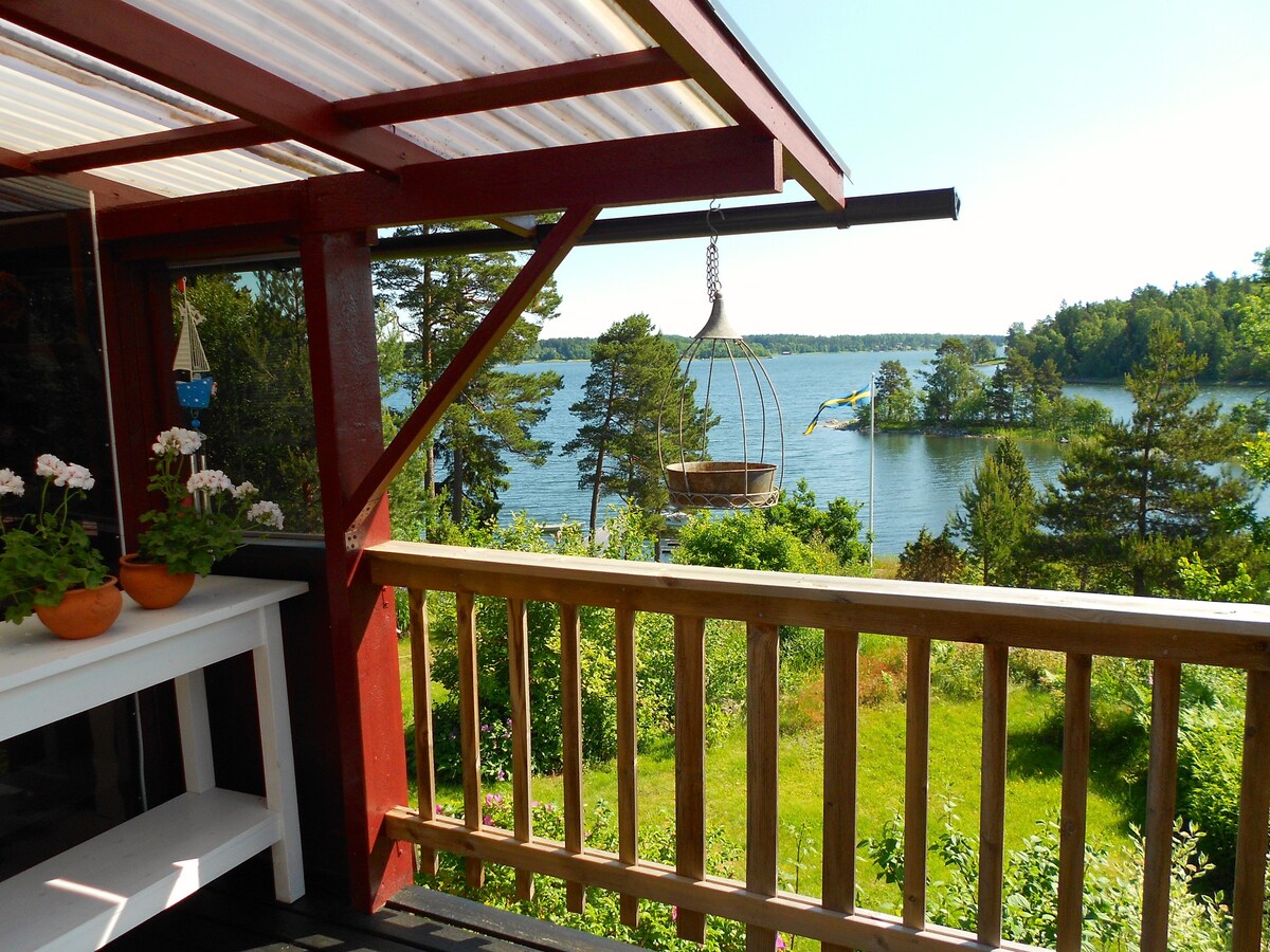 A covered balcony offers a view of the serene waterfront, framed by trees and greenery. A hanging chair adds a cozy touch, complemented by potted flowers nearby. The scene captures a peaceful outdoor space perfect for relaxation.