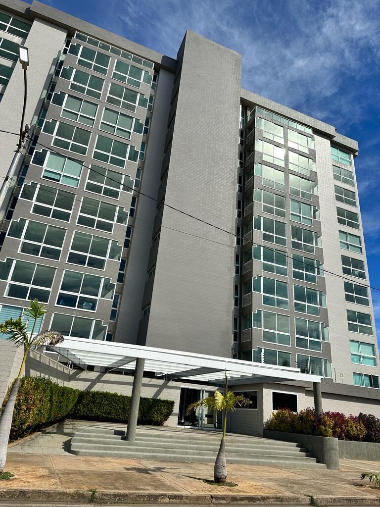 The exterior of a modern multi-story building is displayed, featuring large glass windows and a sleek gray facade. A covered entrance with a small overhang is visible, complemented by landscaped greenery along the front, creating a welcoming first impression.