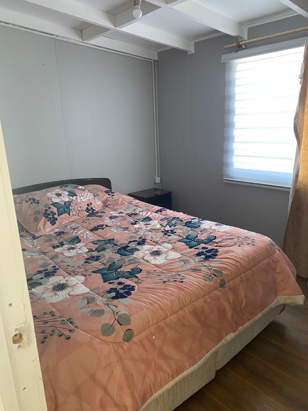 A cozy bedroom features a spacious bed with a floral-patterned comforter in soft pink hues. Natural light filters through a sheer window treatment, illuminating the warm wooden floor. A nightstand is positioned beside the bed, enhancing the room's functionality.