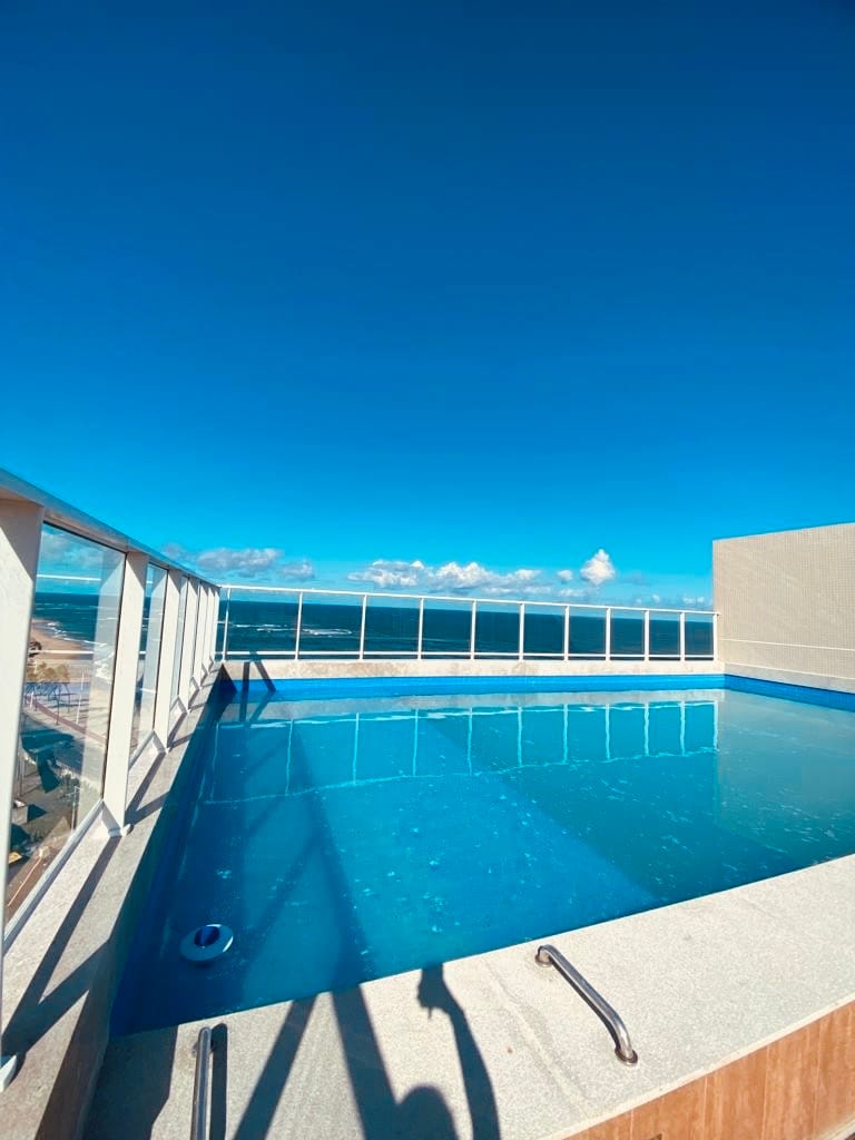 An outdoor swimming pool is situated on a rooftop, surrounded by glass railings. The clear blue water reflects the bright sky, while distant clouds add depth to the serene atmosphere. The scene conveys a sense of openness and relaxation in a spacious setting.
