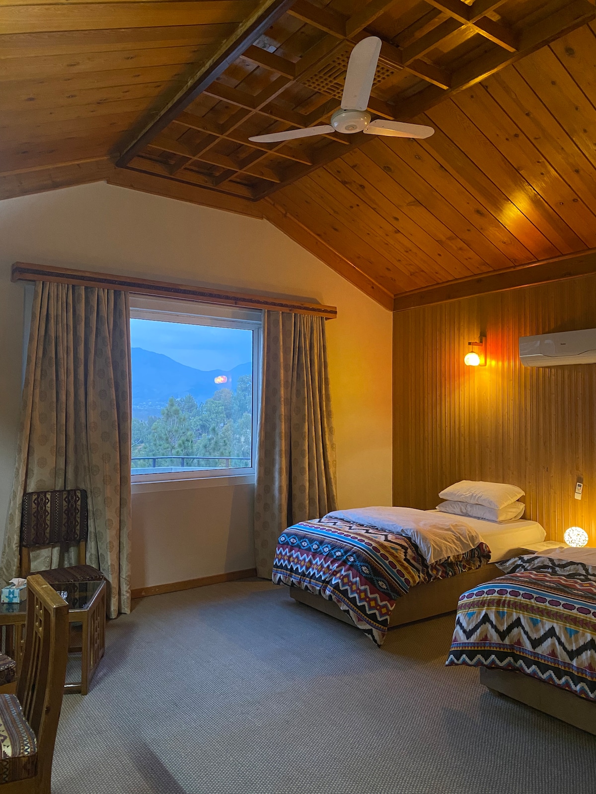 A cozy bedroom features two single beds adorned with patterned blankets. Large windows offer views of the mountains, while warm lighting is provided by wall fixtures. A wooden ceiling enhances the rustic charm of the space. A small table and chairs are situated in the corner.