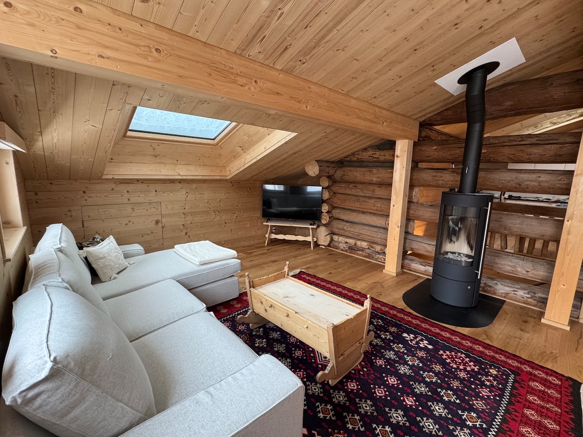 A spacious living area features a light-colored sectional sofa arranged around a wooden coffee table. A flat-screen television is mounted on a wall, complemented by a modern black stove, which adds a touch of warmth. Natural light streams through a skylight overhead.