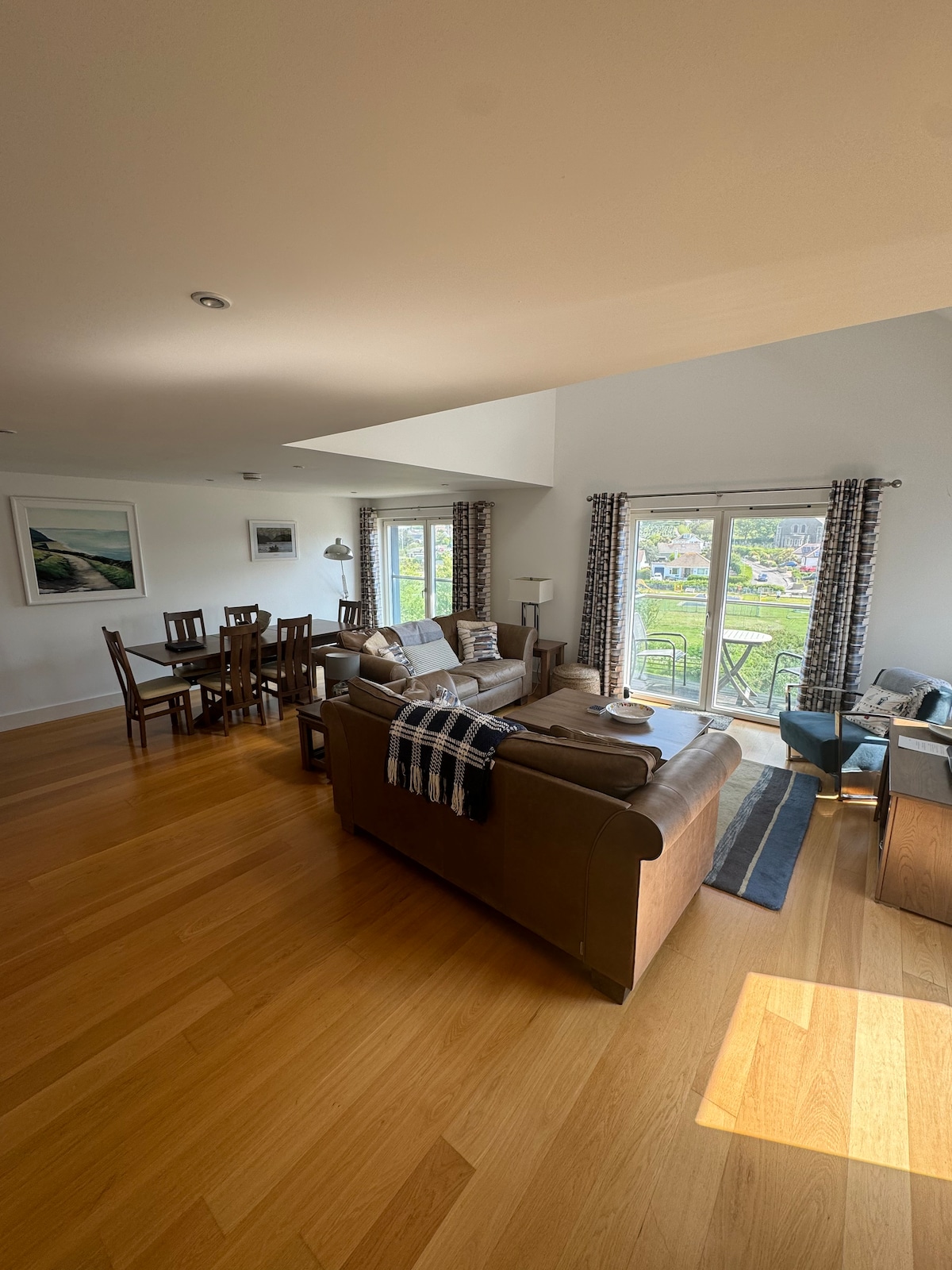 A spacious living area features modern seating with soft cushions and a coffee table. A dining area with wooden chairs is visible, illuminated by natural light streaming through large windows. French doors open to a balcony, offering views of the surrounding outdoors.