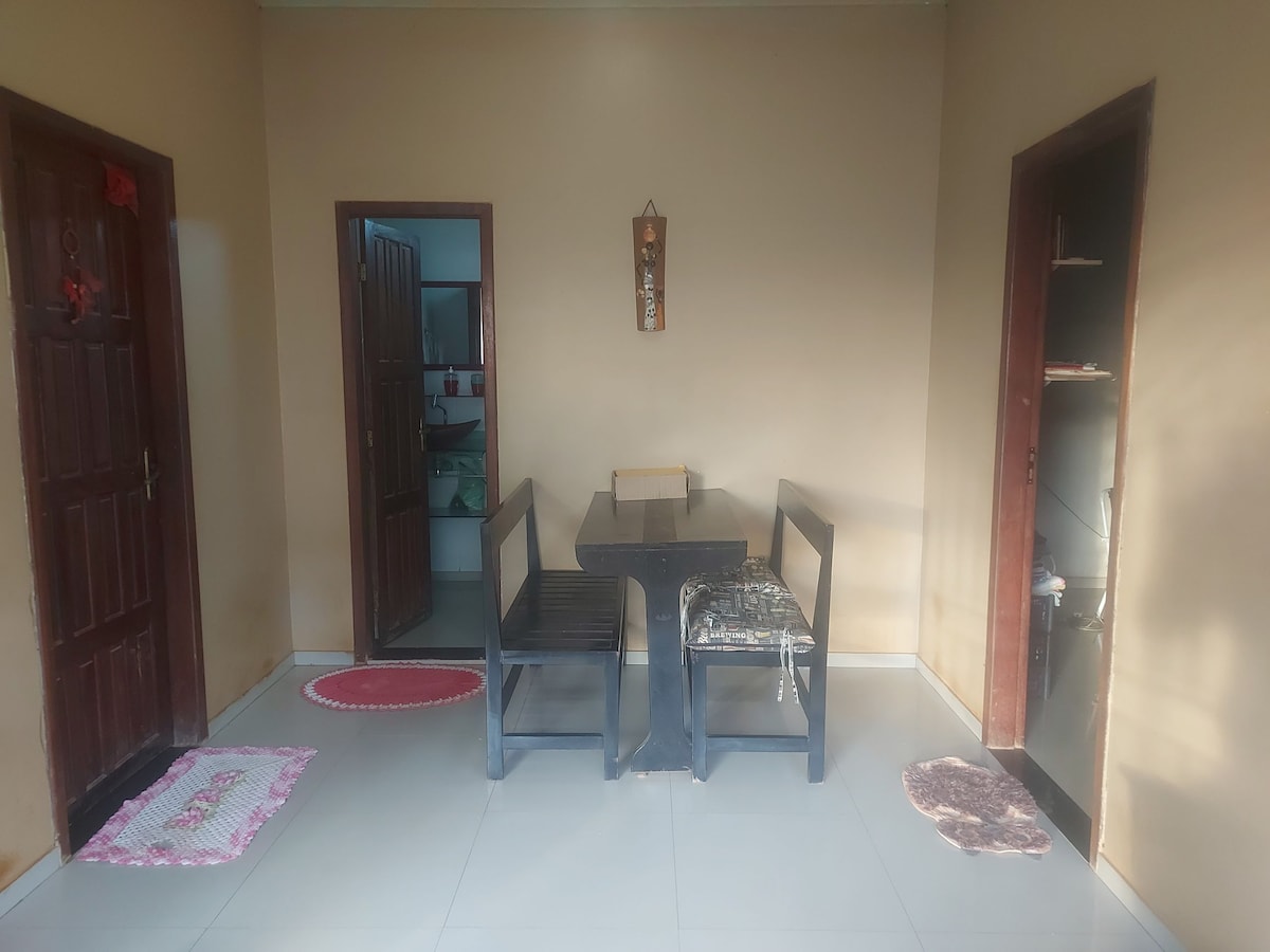 A simple dining area features a black dining table surrounded by two wooden chairs. The neutral-toned walls are complemented by two doors, leading to other rooms. Small mats are placed on the tiled floor near the table, adding a touch of warmth to the space.