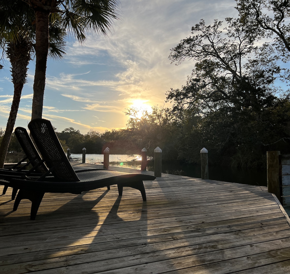 A wooden dock extends over the water, featuring two loungers positioned for relaxation. The setting sun casts a warm glow, illuminating the surrounding trees and river. Palm trees frame the scene, enhancing the tranquil atmosphere.