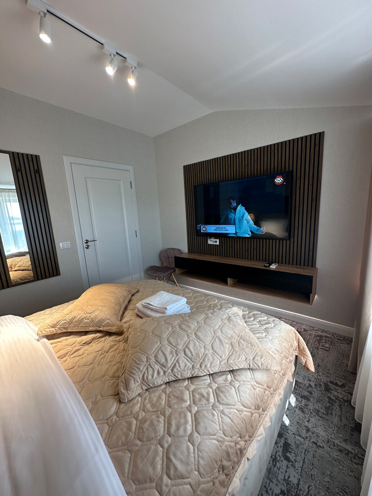 A modern bedroom features a spacious bed with a textured beige quilt and neatly arranged towels. The wall-mounted television is positioned above a sleek wooden console. Mirrors reflect light, enhancing the room's brightness, while a plush chair is placed thoughtfully in the corner.
