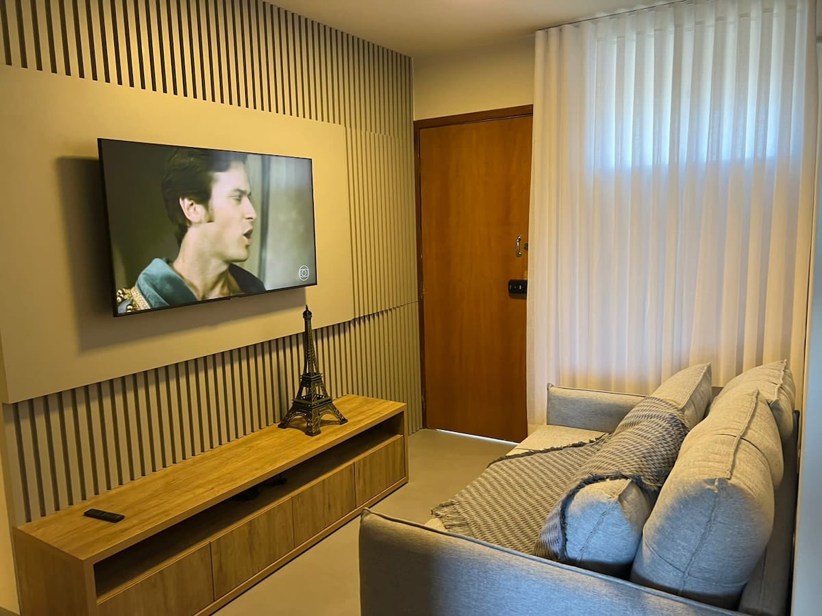 A cozy living area features a light-colored sofa positioned near a wooden entertainment unit. A large television is mounted on the wall, complemented by a decorative Eiffel Tower figurine. Sheer curtains allow natural light to filter in, enhancing the calm ambiance.