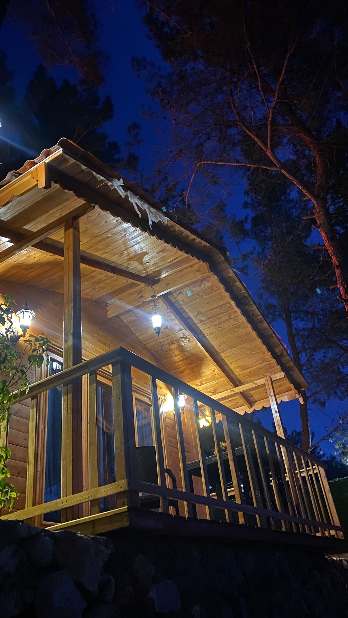 A wooden house is displayed under a starry sky, featuring a spacious covered porch with wooden railings. Illuminated light fixtures provide a warm glow, highlighting the natural wood texture and the surrounding tall trees.