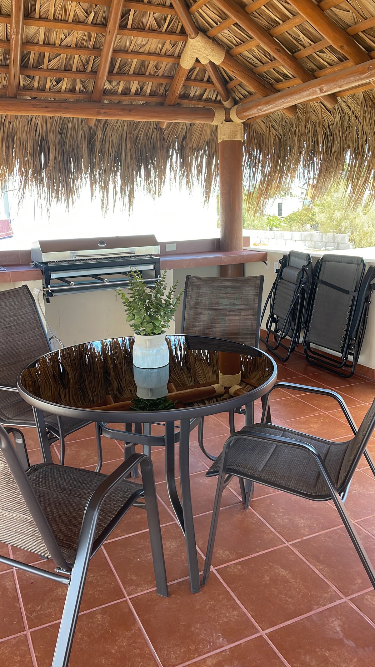 An outdoor dining area features a round table surrounded by four chairs, under a grass-covered palapa roof. A small potted plant sits at the center of the table. In the background, a charcoal grill is visible, with additional folding chairs neatly stacked nearby.