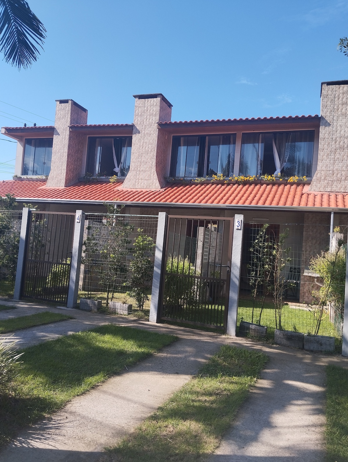 The exterior of a two-story house is displayed, featuring a tiled roof and large windows adorned with sheer curtains. A manicured lawn with greenery surrounds the property, and a gated entrance provides access to a front patio area.