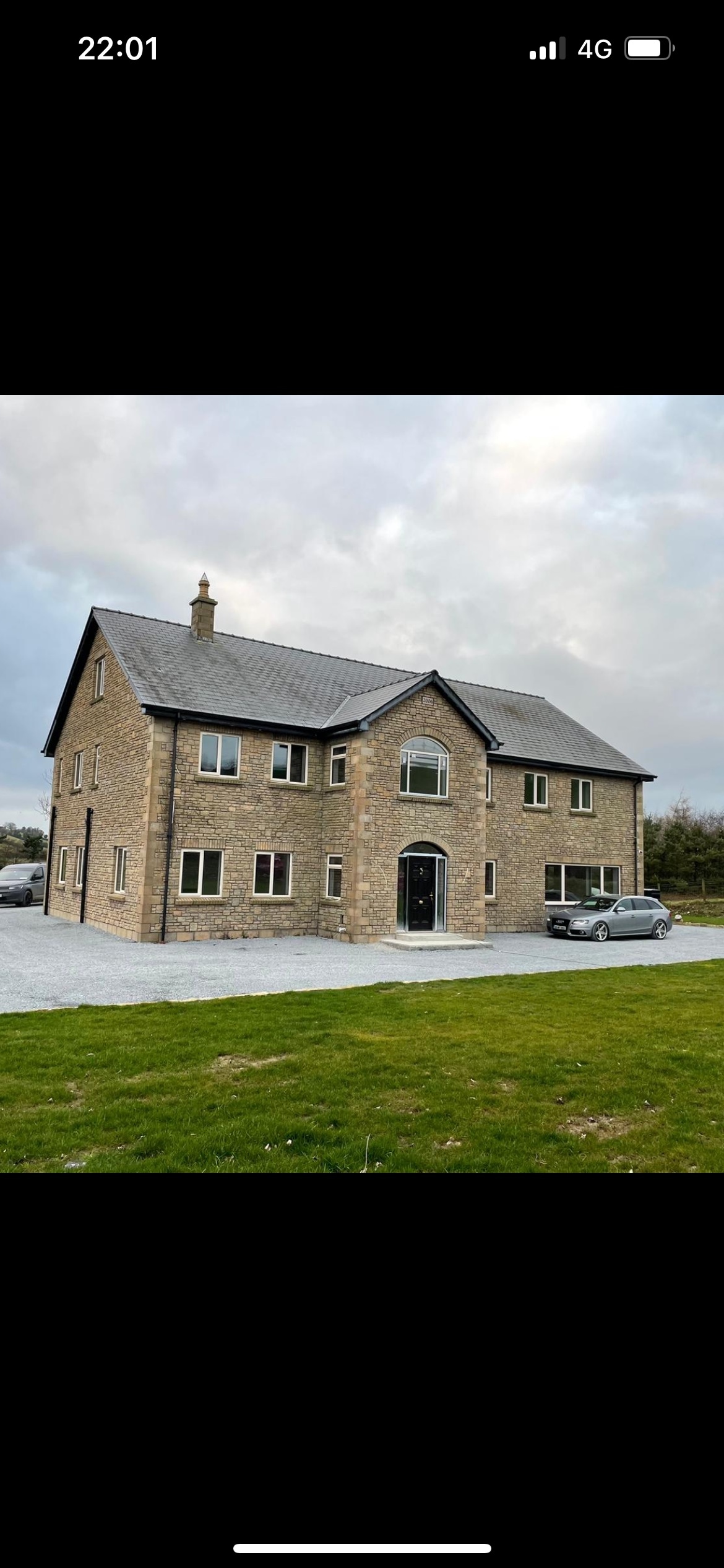 A modern stone house features a two-story design with a grey roof and multiple large windows. A spacious driveway accommodates vehicles, while a well-maintained lawn surrounds the property. The entrance is highlighted by an arched doorframe, inviting guests into the expansive home.