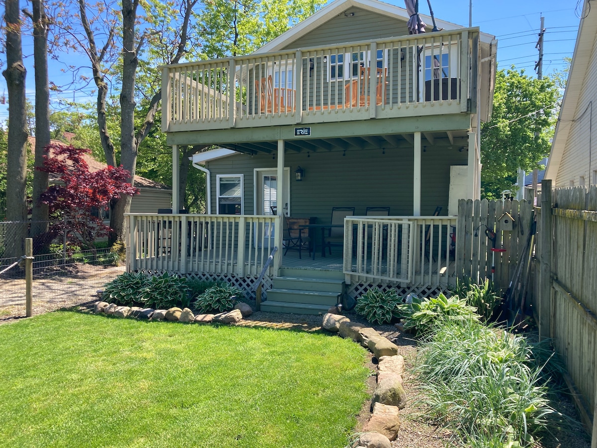 The exterior of the home features a two-story deck with seating arrangements and a well-maintained yard. Lush greenery surrounds the property, including a stone-lined path that leads to the entrance. The inviting facade is complemented by large windows that allow natural light to enter.