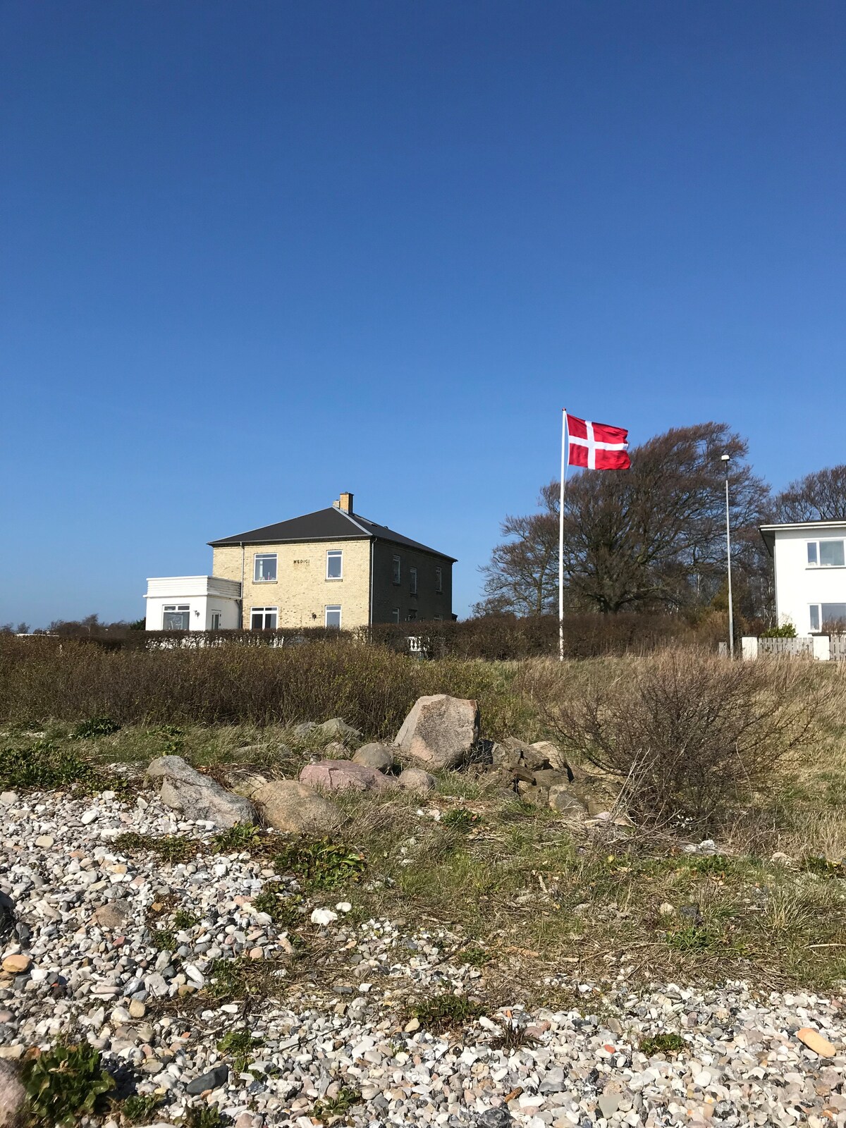 A historic, two-story building is visible among a natural landscape, surrounded by grass and scattered stones. A flag featuring Denmark's national colors is hoisted nearby, contrasting against the clear blue sky. Trees are seen in the background, adding to the serene outdoor setting.