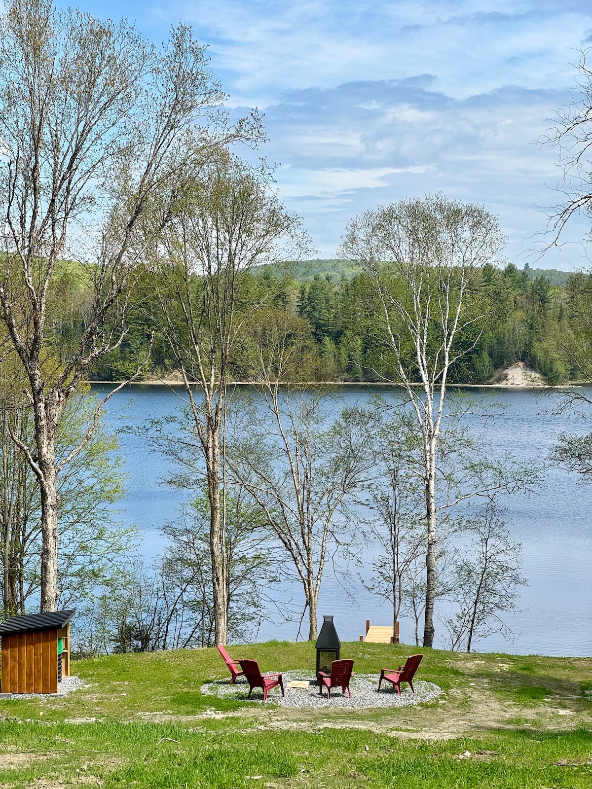 A serene outdoor space is shown, featuring two red chairs positioned around a stone fire pit. The setting overlooks a calm body of water, surrounded by verdant trees and a gently sloping hillside. A small shed is visible in the background.
