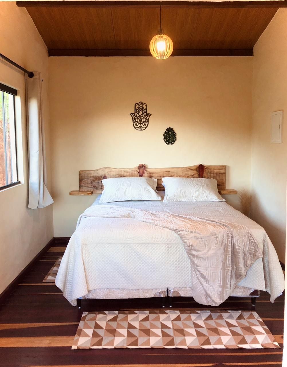 The bedroom features a wooden bed frame with two spacious beds adorned with light bedding. Natural light enters through a large window, complemented by a decorative wall piece and a pendant light hanging from the wood-paneled ceiling.