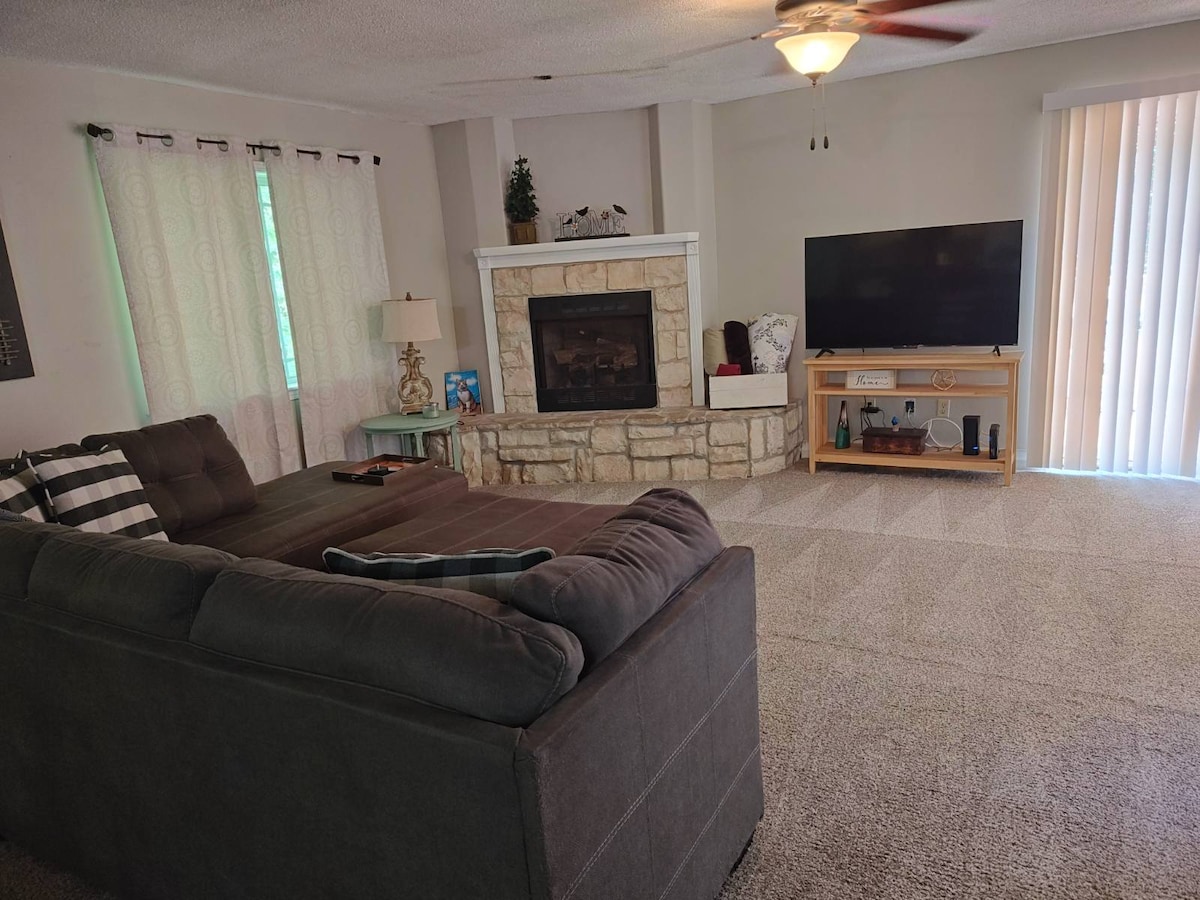 A spacious living area is highlighted by a large sectional sofa and a flat-screen television on a wooden stand. Light streams in through vertical blinds, and a stone fireplace serves as a focal point. Soft carpeting covers the floor, creating a cozy ambiance.