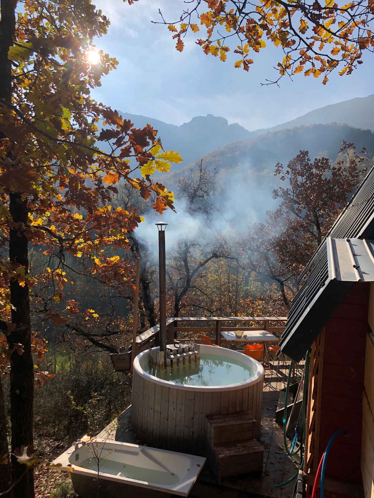 A wooden Nordic bath is situated outdoors, surrounded by autumn foliage and the soft glow of sunlight. Smoke rises gently from a chimney, while distant mountains provide a serene backdrop. A small white tub can be seen nearby, inviting a unique bathing experience.