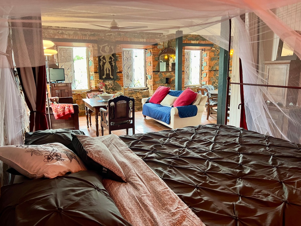 A spacious bedroom is highlighted by a large bed draped with a delicate canopy. The room features exposed stone walls, a sitting area with a colorful couch, and a dining table nearby. Sunlight filters through multiple windows, enhancing the inviting atmosphere.