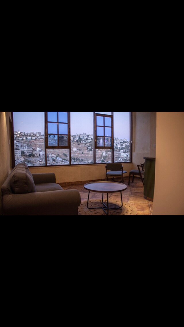 A cozy sitting area features a grey sofa and a small circular table, positioned near large windows that provide views of the surrounding landscape. Natural light fills the space, and a single chair complements the seating arrangement.