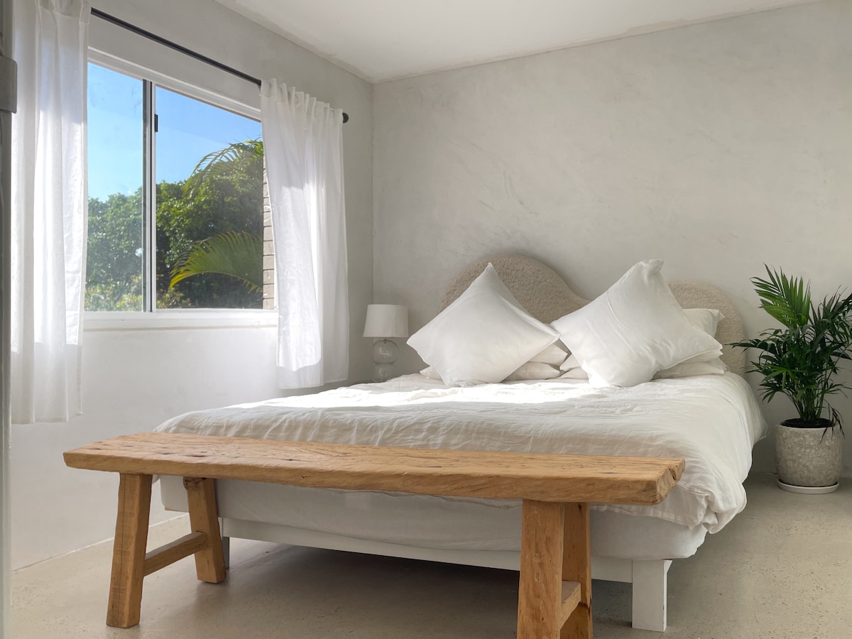 A serene bedroom is featured, with soft natural light illuminating the space through a large window. A spacious bed is adorned with plush pillows, while a wooden bench adds a rustic touch. A small potted plant enhances the calming atmosphere.