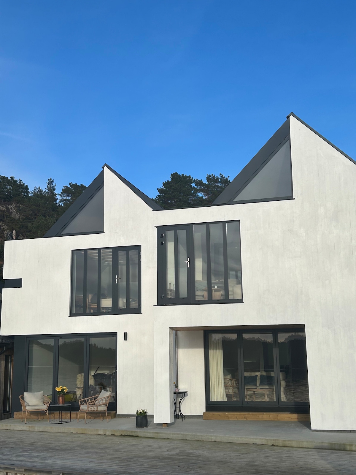 The exterior of a modern two-story house is showcased, featuring a clean white facade and distinctive sloped roofs. Large glass windows provide a view into the well-lit interior, surrounded by trees and a clear blue sky.
