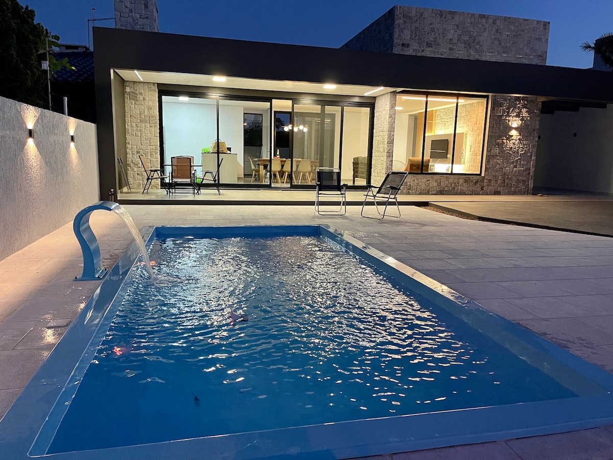 An illuminated swimming pool is visible in the foreground, surrounded by a light-colored patio. The house, with large glass doors, showcases a spacious living area. The exterior lighting enhances the modern design of the home during the evening hours.