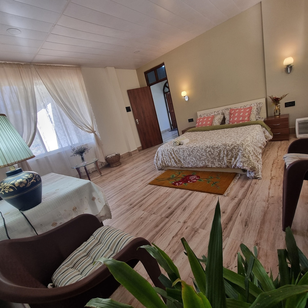 A spacious bedroom features a cozy bed dressed in light linens, with decorative pillows adding subtle color. Natural light filters through sheer curtains, illuminating the wooden floors. A small table with a lamp and two comfortable chairs are arranged nearby, creating a welcoming space.