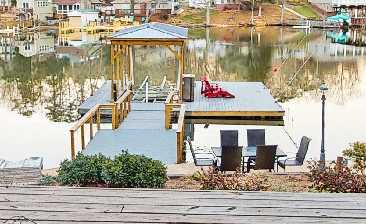 A lakeside dock with a covered seating area features a set of six dark chairs surrounding a table. A red lounge chair is positioned nearby. The calm water reflects the surrounding homes and trees, creating a serene atmosphere.