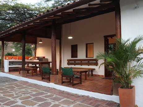 A spacious outdoor terrace features a wooden ceiling supported by large beams. A dining table with multiple seats is visible alongside a separate seating area. Potted plants offer a touch of greenery, while the tiled floor complements the rustic charm of the space.