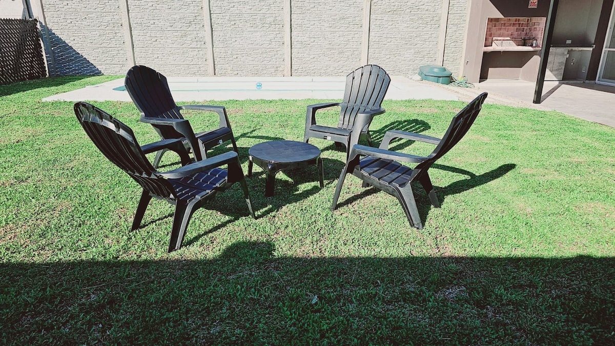 A grassy outdoor space features a circular arrangement of four black chairs surrounding a small table. A swimming pool is visible in the background, complementing the serene setting. The area is shaded, providing a comfortable atmosphere for relaxation.