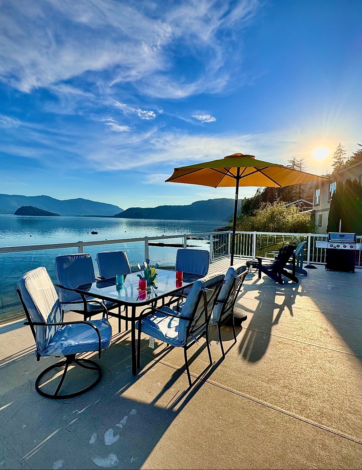 A spacious outdoor deck is shown, furnished with a circular dining table surrounded by six cushioned chairs. An umbrella provides shade over the table, which holds drinks in vibrant colors. The sun sets behind distant mountains, casting a warm glow over the serene lake.