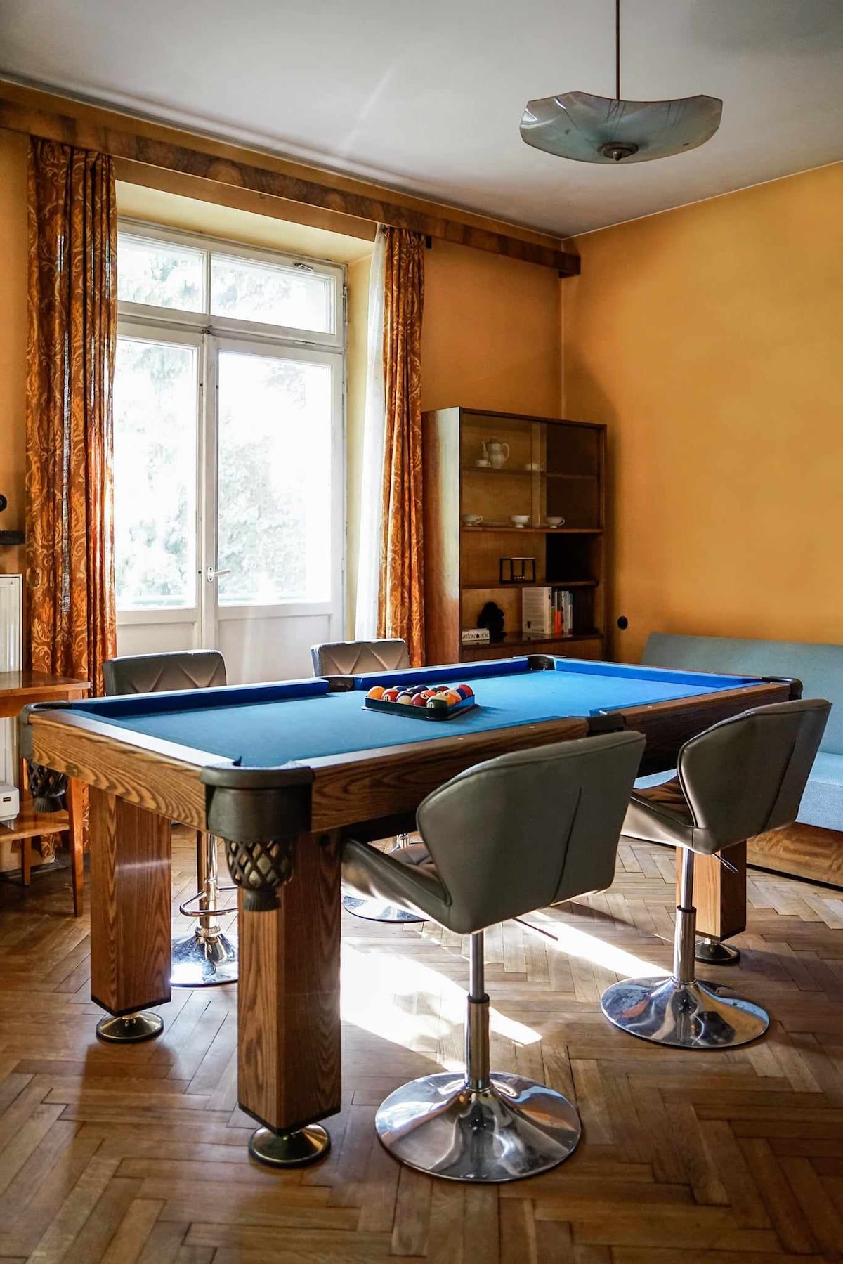 A game room features a blue felt pool table with a complete set of billiard balls arranged on its surface. Surrounding the table, four modern bar stools provide seating. Natural light enters through a large window, complemented by orange curtains that add warmth to the space.