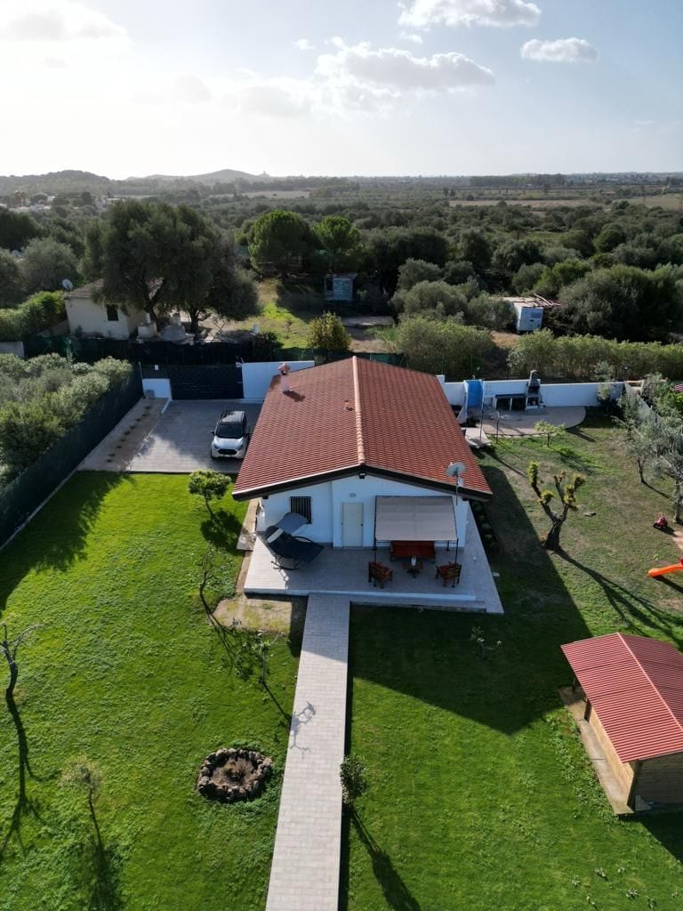 An aerial view of the villa showcases a spacious green lawn surrounding the single-story building. A stone pathway leads to a patio area with outdoor seating. Surrounding vegetation includes trees and shrubs, contributing to a serene, natural environment.