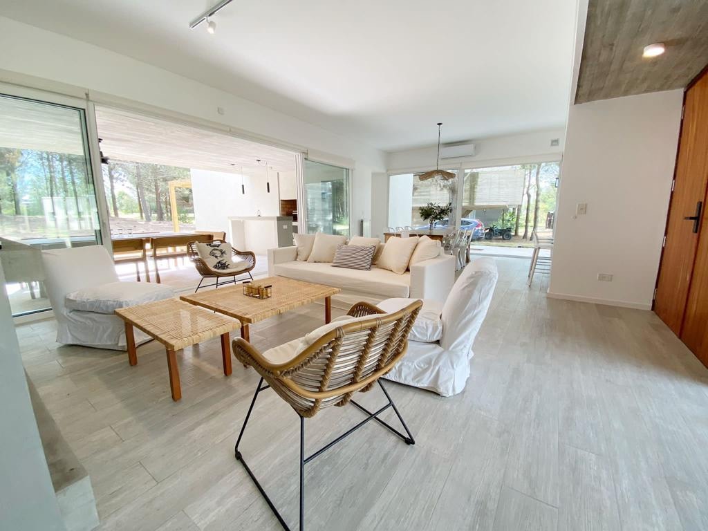 A spacious living area features a light-colored sofa with coordinating pillows, complemented by a wooden coffee table and woven seating. Large glass doors provide a clear view of the outdoors, allowing natural light to fill the room. The open layout connects seamlessly to the dining area.