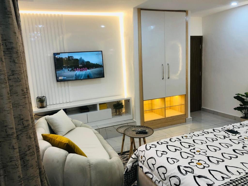 The studio features a comfortable seating area with a light-colored sofa and a side table. A sleek wall-mounted television is positioned above a white console. A spacious bed with a patterned blanket is situated nearby, complemented by a decorative plant, enhancing the cozy ambiance.