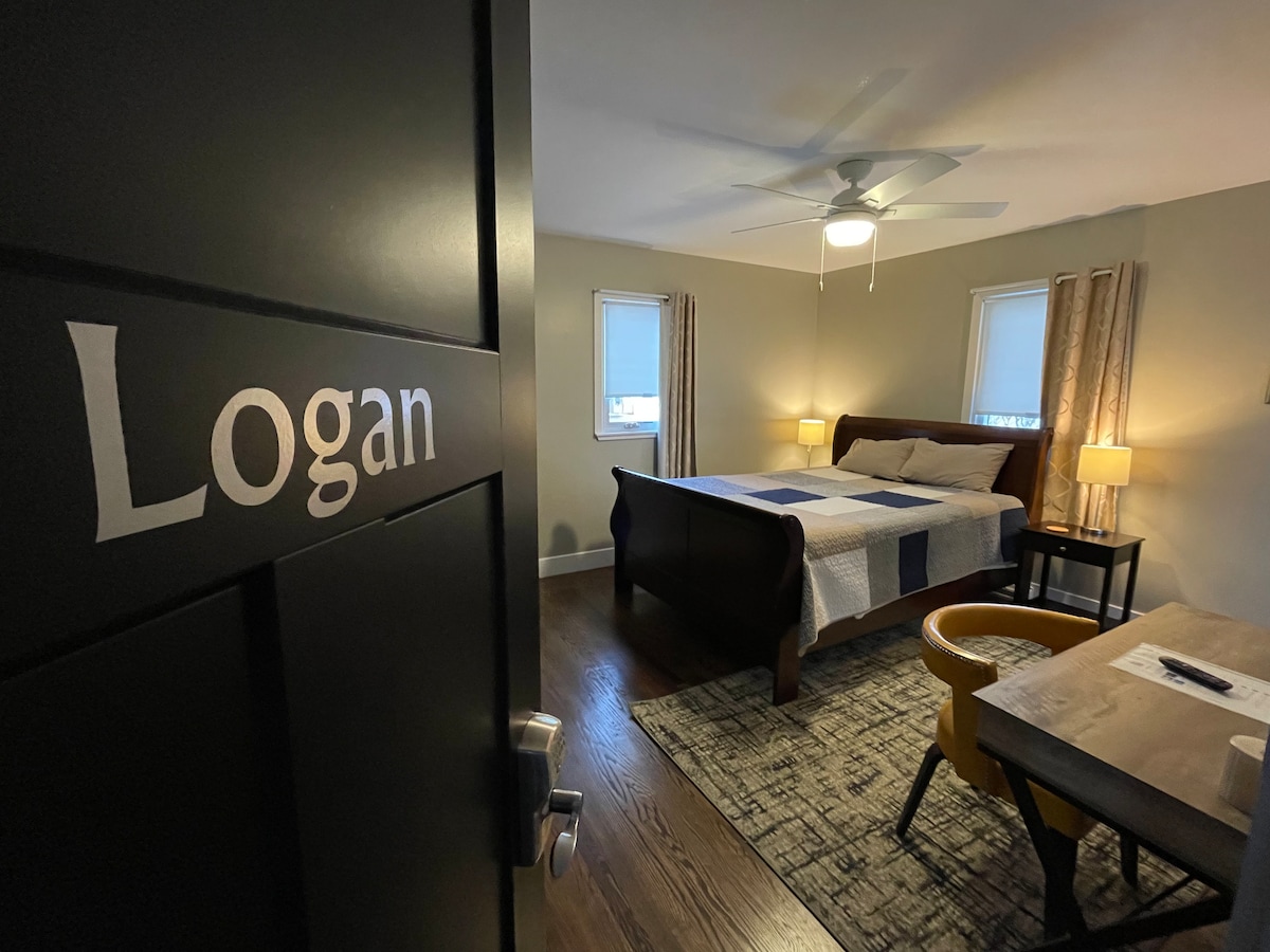 Vue sur la chambre Logan avec lit Queen et deux tables de chevet avec lampes. Bureau avec chaise de restaurant vintage. Les fenêtres ont des stores filtrant la lumière avec des rideaux occultants. 