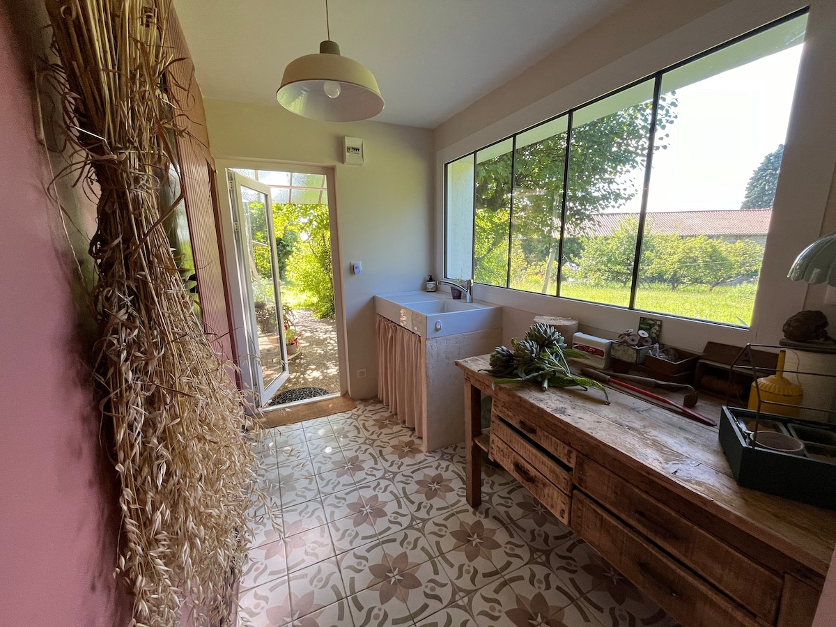 A spacious kitchen area features large windows that provide abundant natural light and views of the garden. A rustic wooden table sits prominently, complemented by light-colored cabinetry. The tiled floor showcases a floral design, adding a touch of charm to the functional space.
