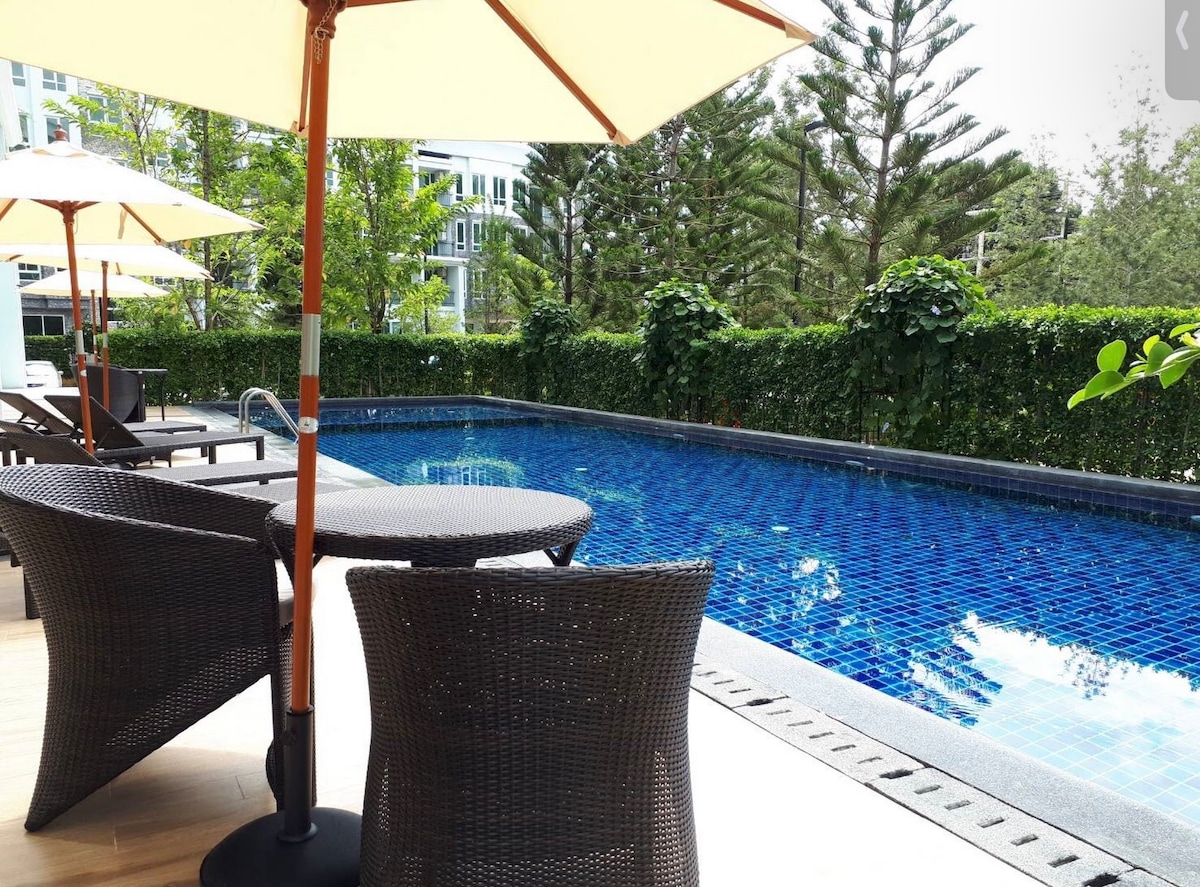An inviting outdoor pool area is set amid lush greenery, featuring a clear blue pool surface and sleek lounge chairs. A round table with umbrella provides shade, enhancing the tranquil environment for relaxation.