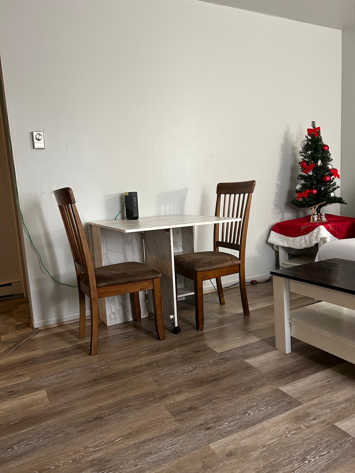 A small dining area is set against a light wall, featuring a compact white table and two wooden chairs. A decorated Christmas tree is positioned nearby, draped with red ornaments and a white skirt, adding a seasonal touch to the space.