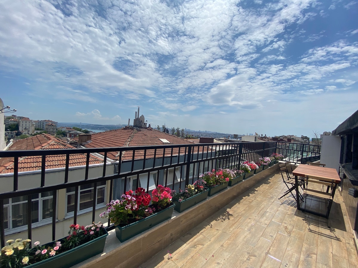 A spacious terrace is adorned with colorful flower boxes overflowing with blooms. An outdoor dining table and chairs set on wooden flooring offer a comfortable space for relaxation. The skyline, with distant views of the sea and rooftops, provides a serene backdrop under a bright, partly cloudy sky.