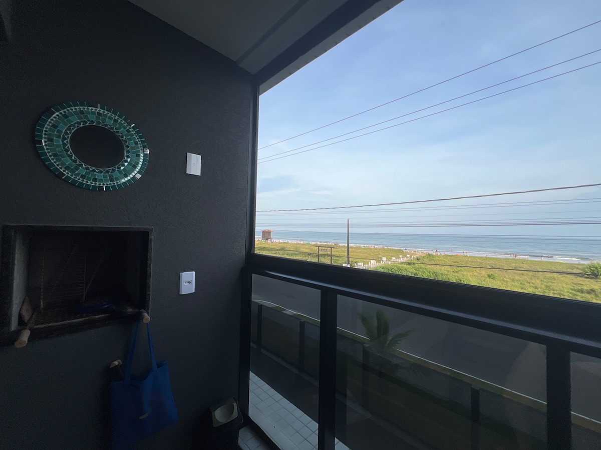 A balcony view showcasing a serene beach scene. The horizon presents the calm ocean, while sun loungers are visible along the sandy shore. Natural light fills the space, highlighting a decorative mirror mounted on the dark wall beside an outdoor cooking area.
