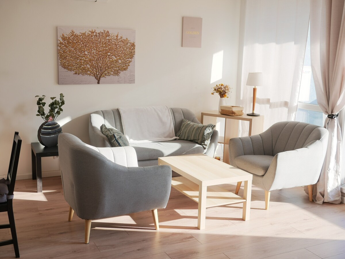 A bright living area features a light-colored sofa with decorative cushions and two comfortable armchairs. A wooden coffee table is centered between the seating. Natural light filters through sheer curtains, creating a warm ambiance. A piece of artwork hangs on the wall, complemented by a small plant.