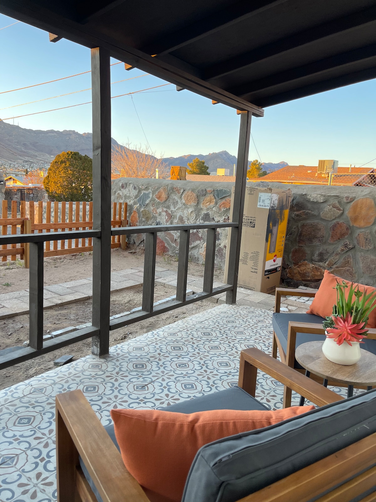 A covered porch features wooden seating with orange pillows and a small round table topped with flowers. The stone walls and surrounding landscape can be seen in the distance, highlighting the scenic view framed by the porch's rails.