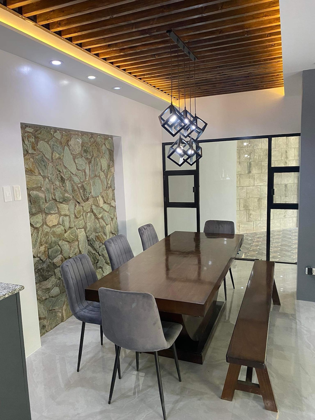 A modern dining area is highlighted by a rectangular wooden table complemented by a wooden bench and four upholstered chairs. The ceiling features a wood panel design, while a unique light fixture hangs above, providing soft illumination. A stone wall adds texture to the space.