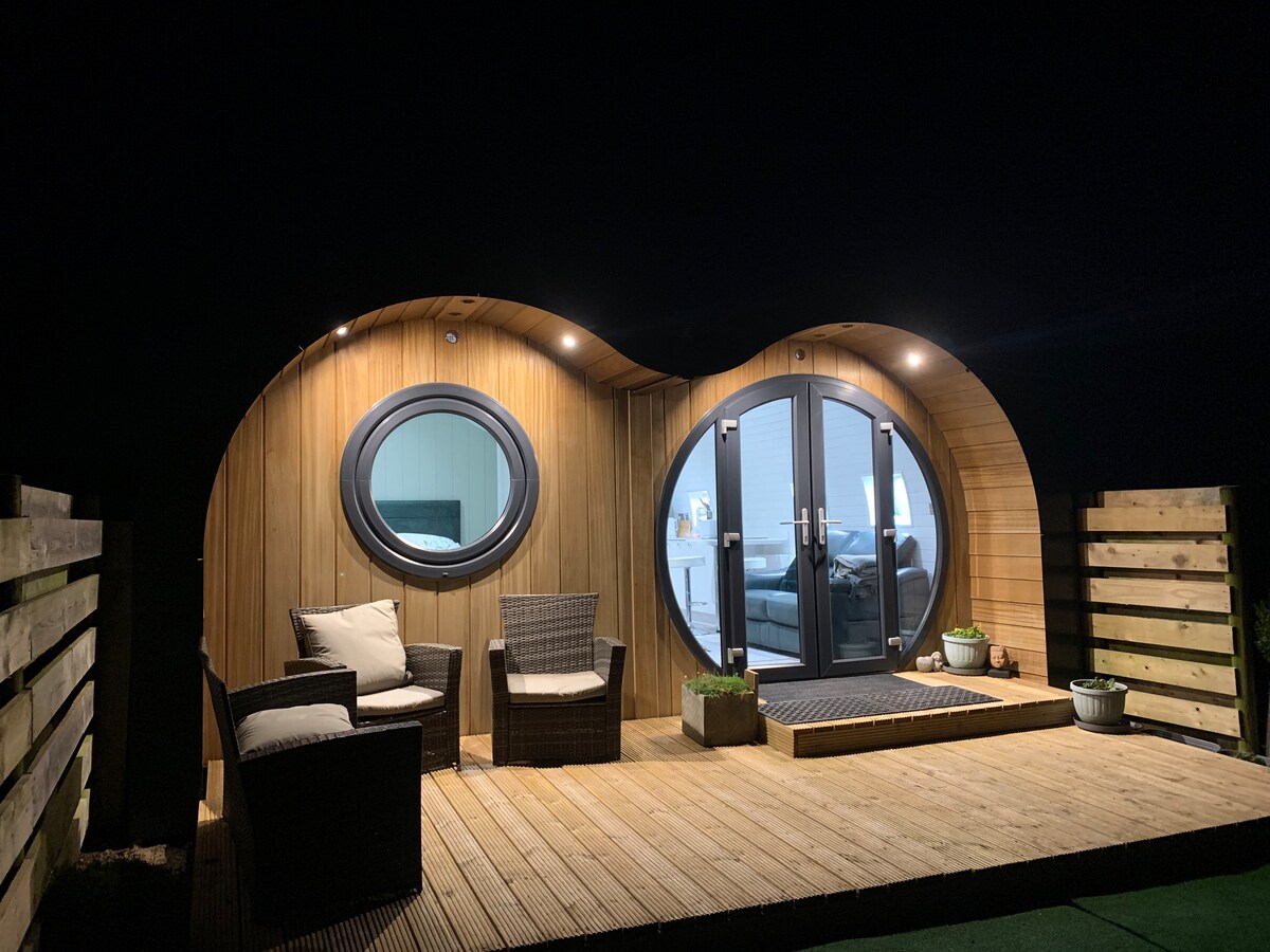 An inviting outdoor deck features two armchairs and a small table beside a rounded entrance with large glass doors. The wooden exterior is complemented by subtle lighting, highlighting the unique pod shape and creating a warm ambiance at night.