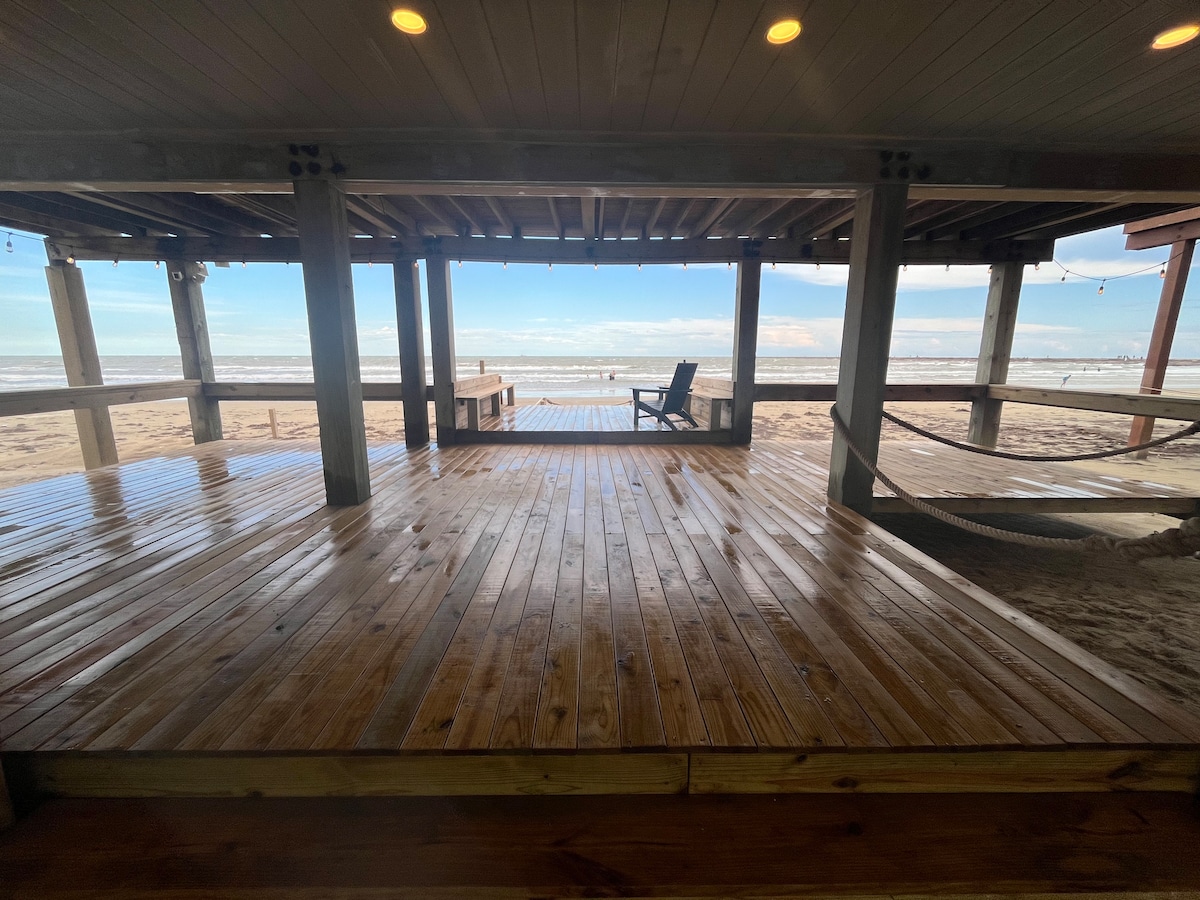 A spacious wooden deck is showcased, offering direct views of the beach and ocean. The expansive area features seating options, including a chair and a hammock, providing ample space for relaxation. Natural light floods the deck, highlighting the inviting coastal atmosphere.