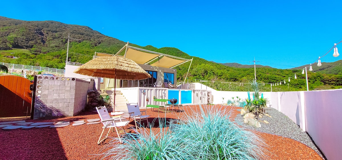 The outdoor area features a spacious layout with a sunshade structure providing shade over a seating arrangement. Lush greenery and gravel accent the surroundings, while a swimming pool sits invitingly in the background, bordered by rustic stone pathways.
