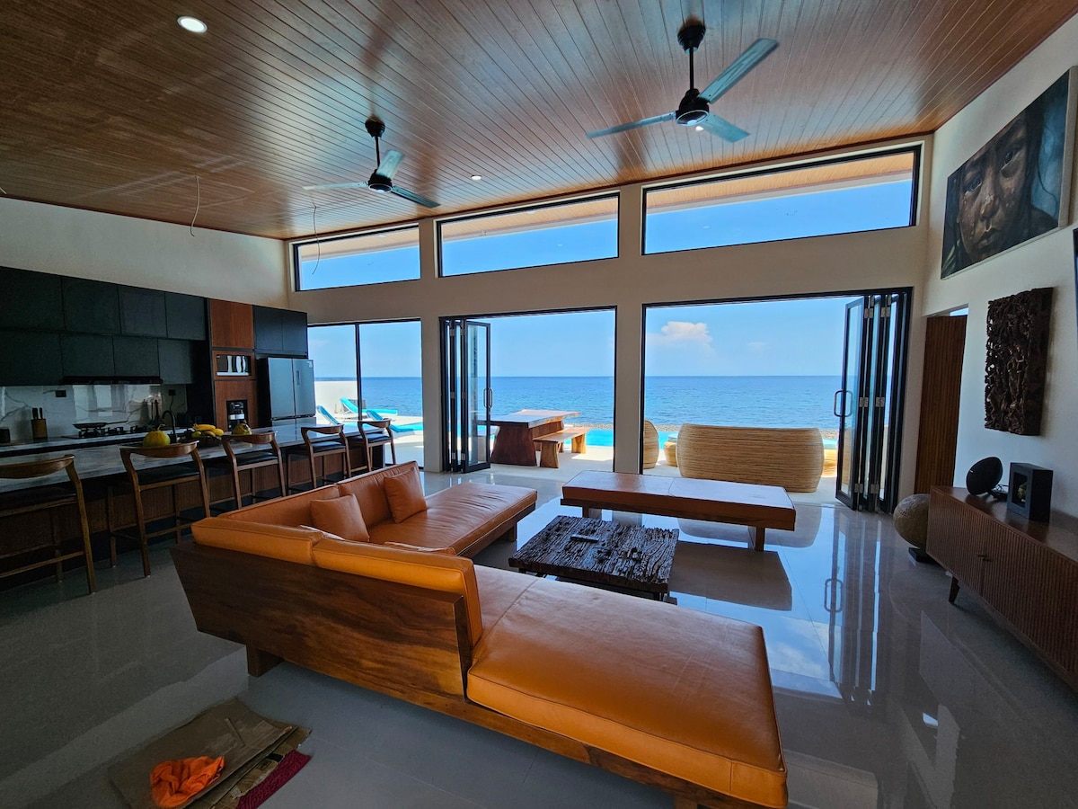 A spacious living area is highlighted by large glass doors, providing stunning sea views. Contemporary furnishings, including a comfortable orange sofa and a wooden coffee table, complement the neutral-toned decor. Natural light floods the space, enhancing the open, airy atmosphere.