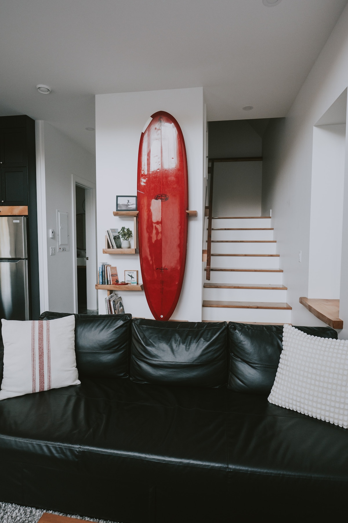 A sleek black couch is positioned in the foreground, adorned with two decorative pillows. A vibrant red surfboard is mounted on the wall behind it, adding a splash of color. Soft natural light illuminates the space, enhancing the open staircase in the background.